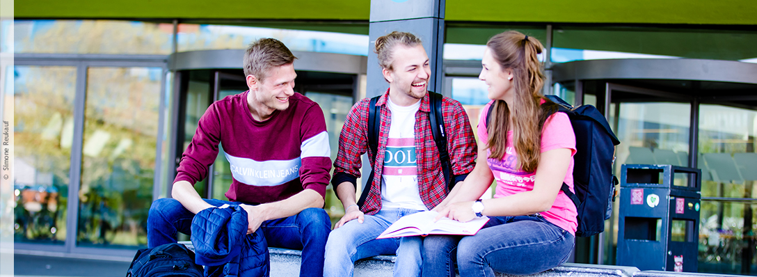 Drei Studierende sitzen gut gelaunt vor einem Gebäude auf dem Campus Westerberg