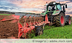 Ein Traktor auf einem Feld mit angespanntem schwerem Gerät pflügt ein Feld um, im Hintergrund sind dunkle Wolken am Himmel